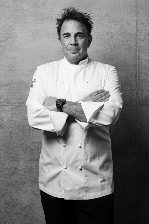 A man wearing a white chef’s coat stands with his arms crossed, looking at the camera, in front of a textured concrete wall. The image is in black and white.