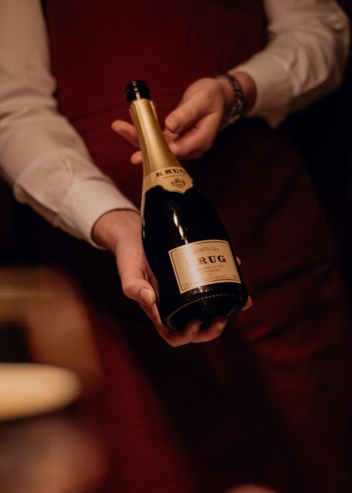 A person wearing a white shirt and red apron holds a bottle of Krug Champagne, presenting it with both hands in a dimly lit setting at the exclusive Krug 4-Hands-Dinner.