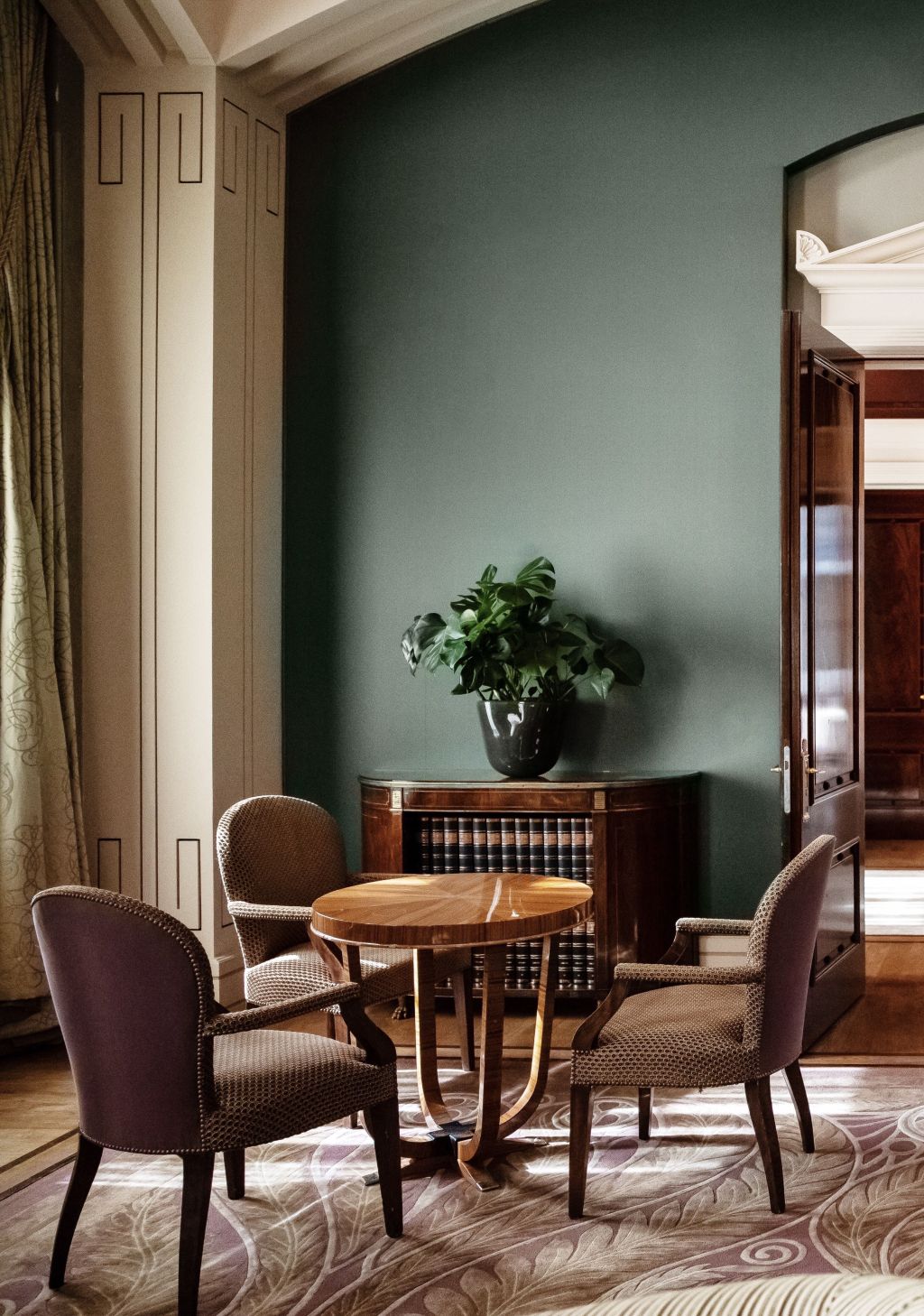 A stylish room with three upholstered chairs around a wooden round table, perfect for afternoon tea. Set on a patterned rug, a green plant sits on a sideboard against a muted green wall, with an open door revealing another room.