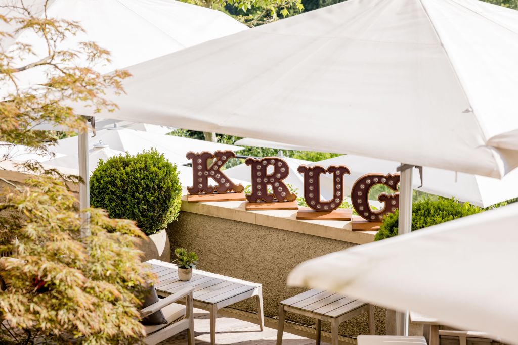 Wooden patio with white umbrellas, small tables, green shrubs, and large marquee letters spelling KRUG placed on a ledge in the background at the Krug Lounge Dolder Grand hotel terrace.