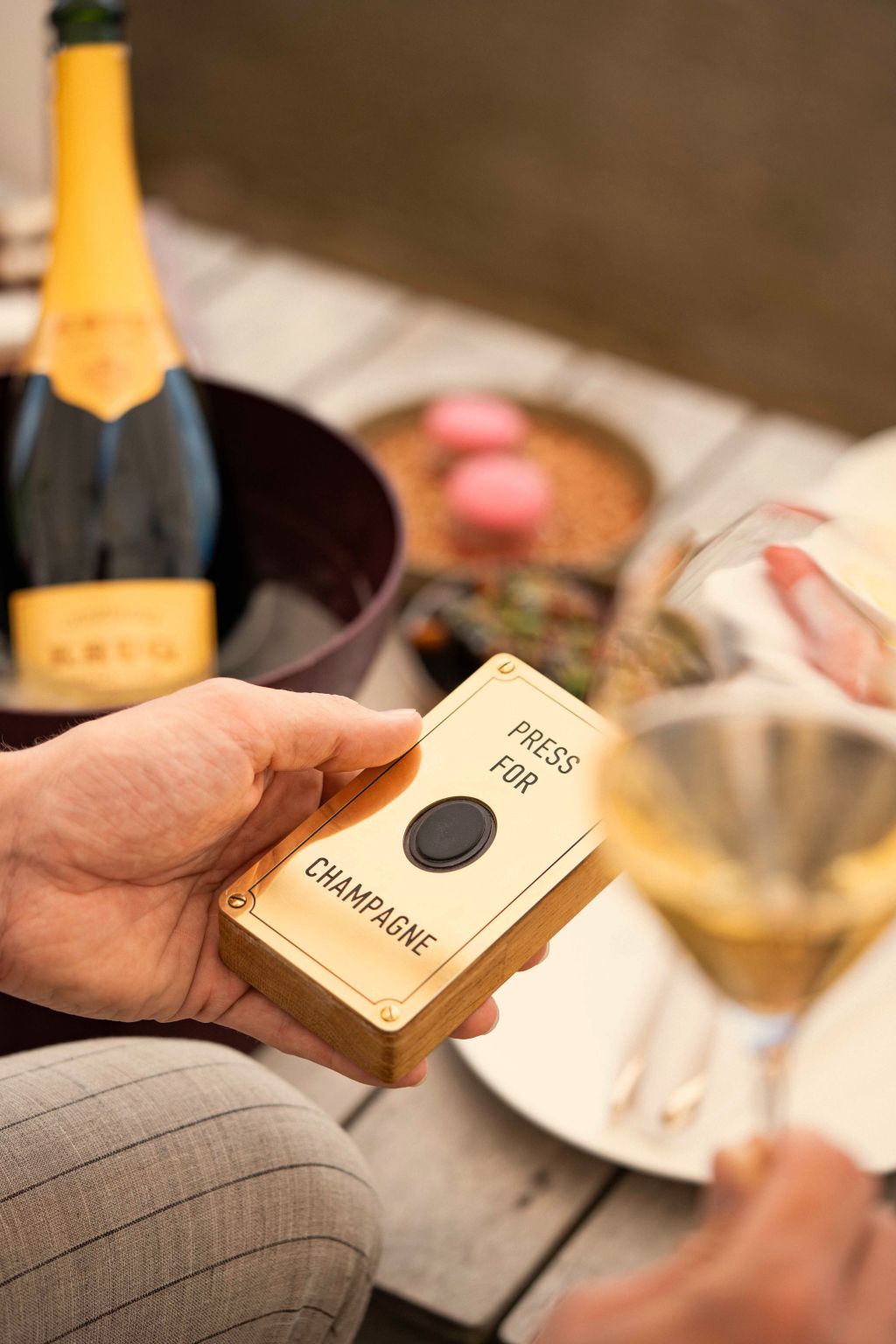 Eine Person hält einen Knopf mit der Aufschrift Press for Champagne und ein Glas Krug Champagner in der Hand. Im Hintergrund stehen eine Flasche Champagner, Snacks und rosa Macarons auf einem Holztisch auf der Terrasse Zürich der Krug Lounge Dolder Grand.