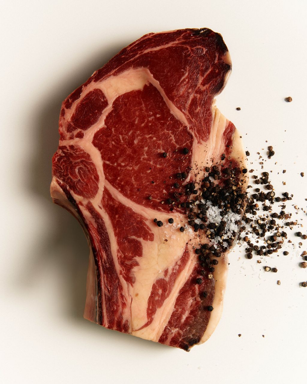 Ein rohes Ribeye-Steak mit sichtbarer Marmorierung ruht auf einer hellen Unterlage, bestreut mit grobem Salz und schwarzem Pfeffer - ein erstklassiges Stück von The Grand Meatery.
