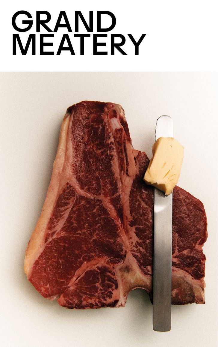 Ein rohes T-Bone-Steak liegt auf einer weißen Fläche, auf der ein Stück Butter auf einem Messer ruht. Darüber steht in fetter schwarzer Schrift GRAND MEATERY.