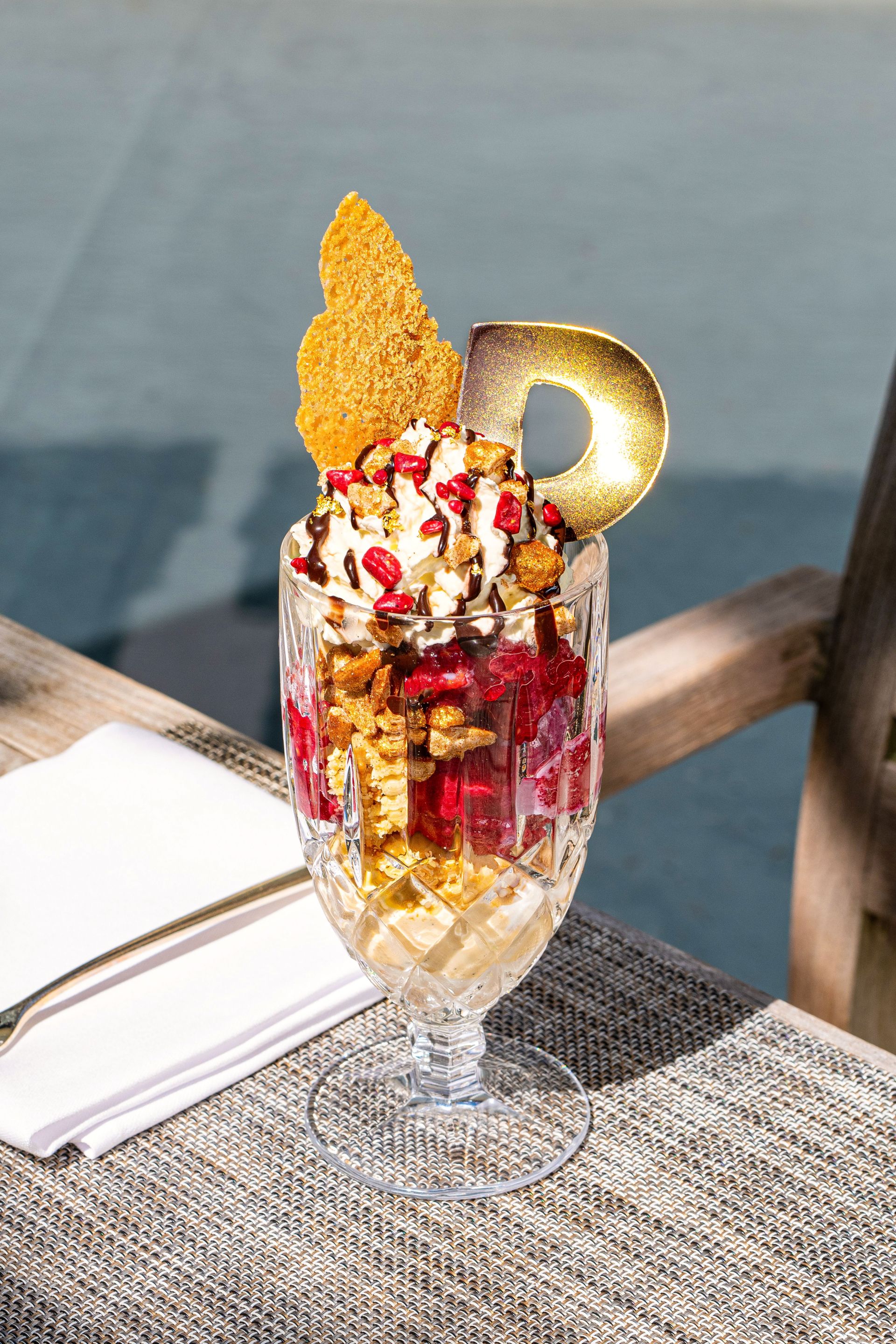 A tall crystal glass filled with a Saltz Ice Creme layered dessert, topped with whipped cream, pomegranate seeds, caramel drizzle, a golden cookie, and a chocolate “D,” sitting on a table with a napkin and fork.
