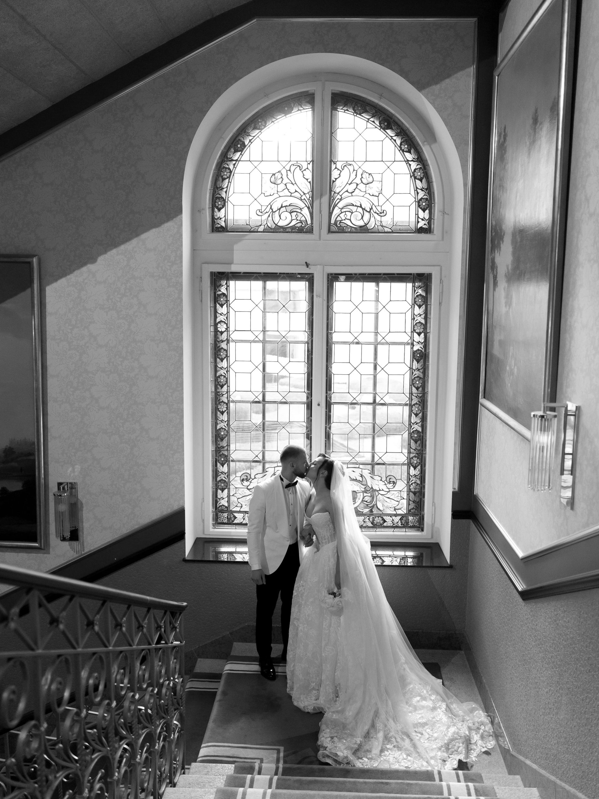 A bride and groom share a kiss at the bottom of a staircase in front of a large, ornate stained glass window. The bride wears a long veil and gown, and the groom is dressed in a suit. The photo is in black and white.