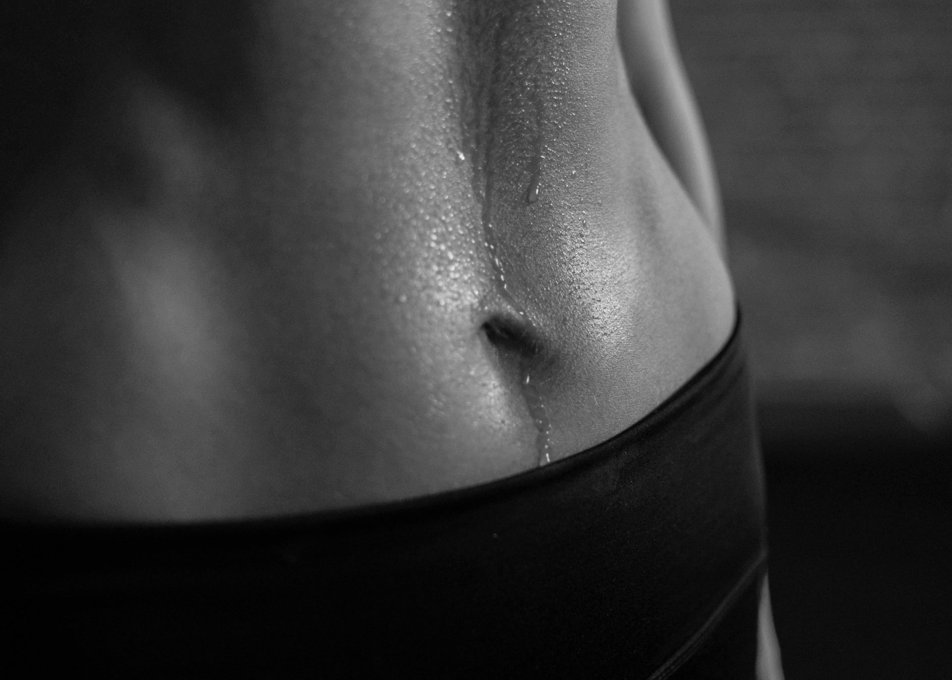 Close-up of a person's sweaty midsection, highlighting defined abs and a trail of sweat dripping from the stomach to the black waistband of athletic shorts. Captured in black and white, this shot evokes the intensity found at Dolder Grand Fitness studio.