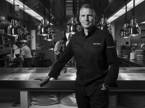 Ein Koch in dunkler Uniform steht selbstbewusst an einer Küchentheke, über der helles Licht hängt. Im Hintergrund arbeiten andere Küchenmitarbeiter, und unter der Theke sind Regale mit Tellern zu sehen, die eine professionelle Küchenumgebung schaffen.