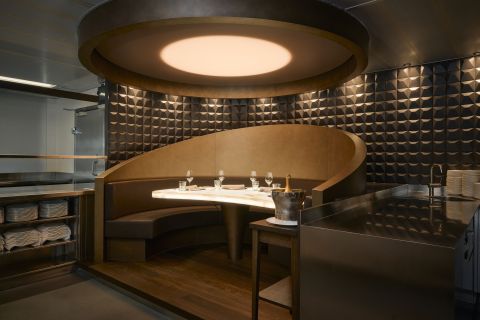 Eine moderne, gehobene Chef's Table-Kabine mit einer geschwungenen braunen Lederbank, einem Marmortisch für vier Personen, einer geometrisch strukturierten Wand und einer weichen, runden Deckenleuchte; daneben stehen auf einer Metallplatte Teller und ein Eiskübel mit einer Flasche.