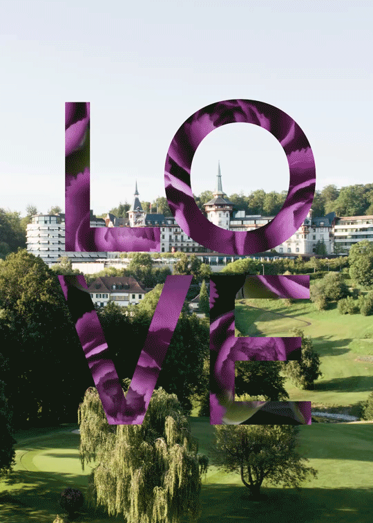 Large letters spelling LOVE, filled with a purple floral texture, stand over a scenic landscape of green lawns and trees. The vibrant display adds an Easter charm to the view, contrasting beautifully with the background buildings.