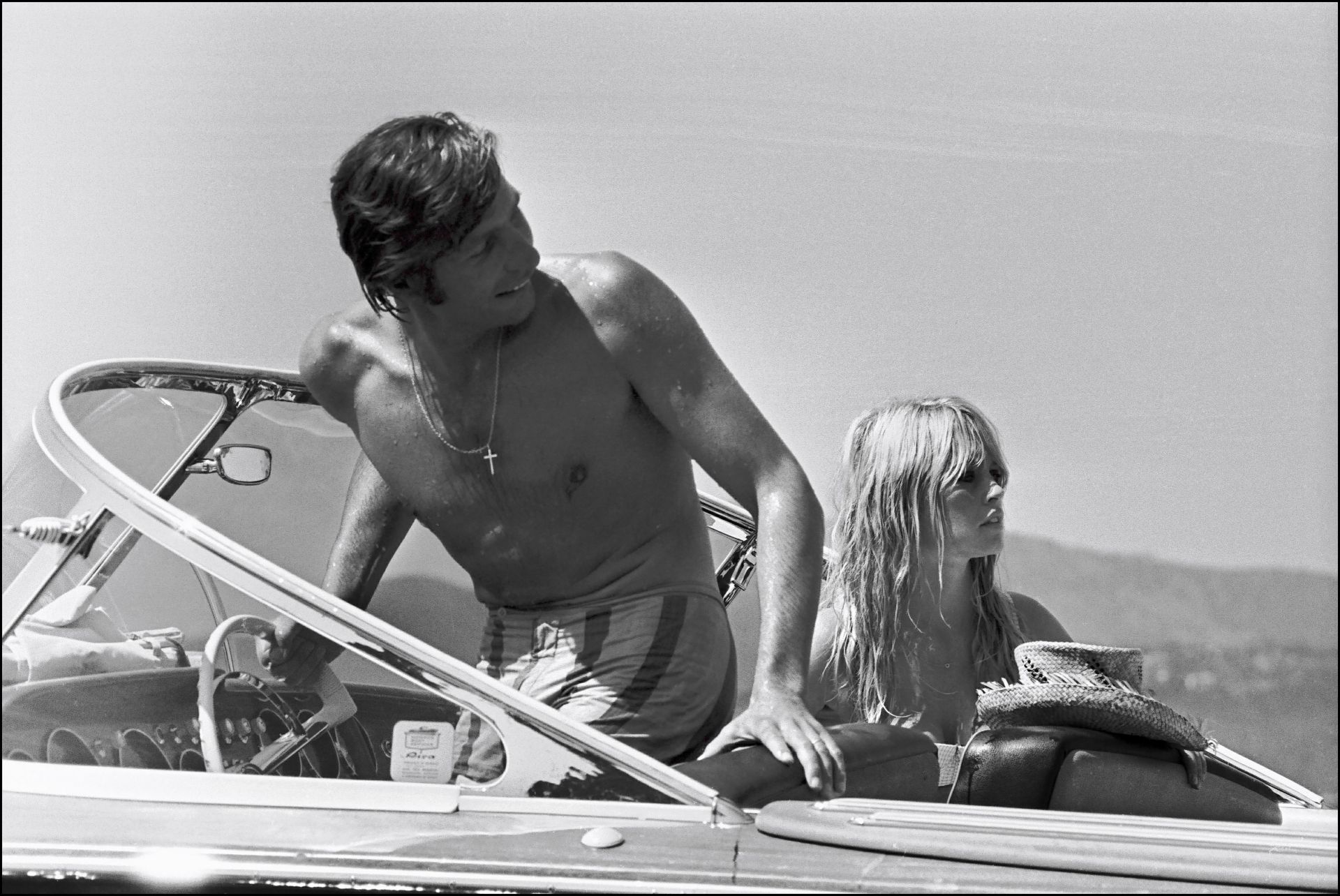 Ein Mann ohne Hemd und eine Frau im Bikini sitzen in einem kleinen Motorboot. Der Mann steht am Steuerrad und schaut zur Seite, während die Frau hinter ihm sitzt und einen Strohhut auf dem Schoß trägt. Im Hintergrund sind Berge und Wasser zu sehen.