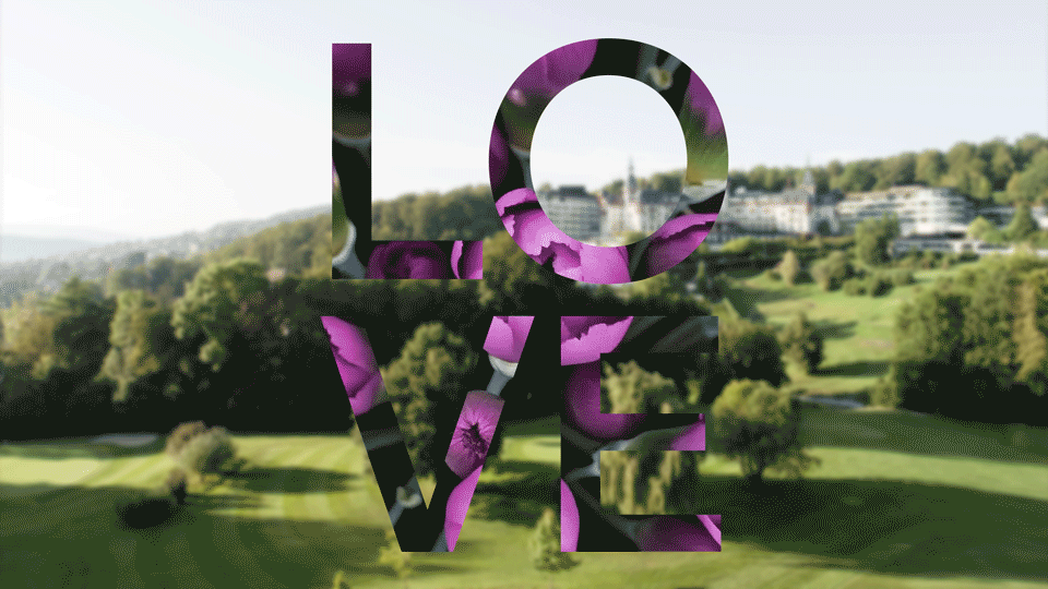 Das Wort LOVE in fetten Großbuchstaben, gefüllt mit einem lila Blumenmuster, überlagert von einem verschwommenen, an Ostern erinnernden Hintergrund einer grünen, sonnigen Landschaft mit Bäumen und Gebäuden.