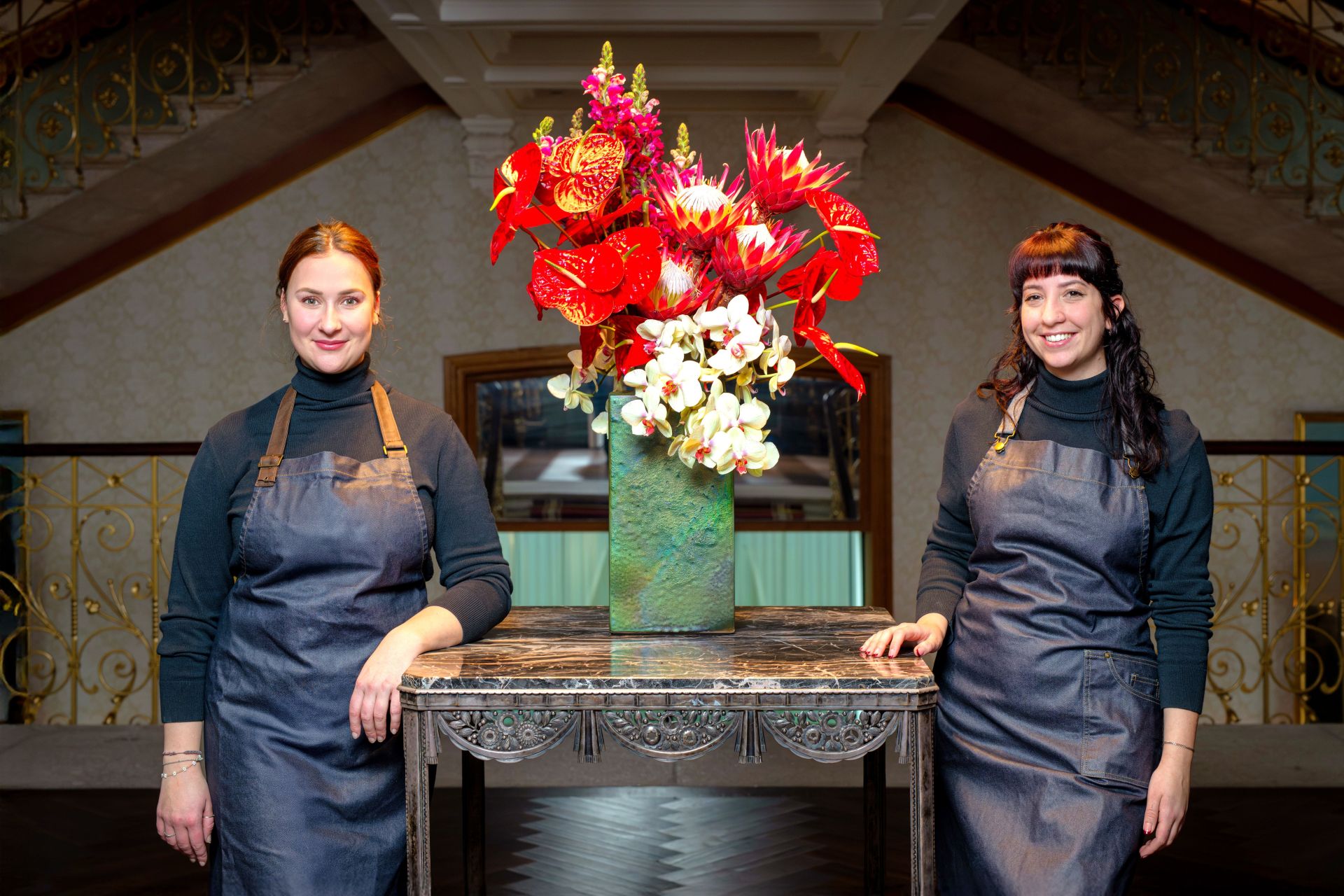 Two women wearing dark aprons stand on either side of a table with an ornate vase full of vibrant red and white flowers. They are indoors, in a well-lit, elegant space with gold railings and patterned walls.