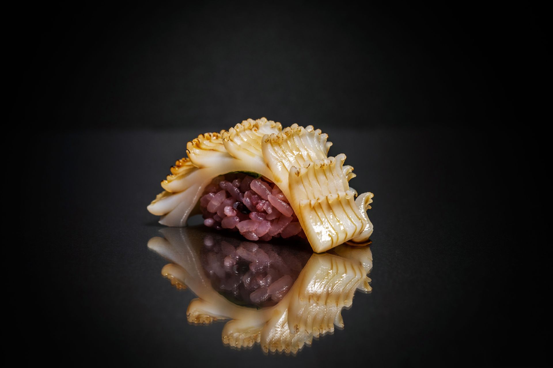A single piece of sushi with a ridged white topping and purple rice is displayed on a shiny, dark surface at a renowned omakase restaurant, reflecting its image against a black background.