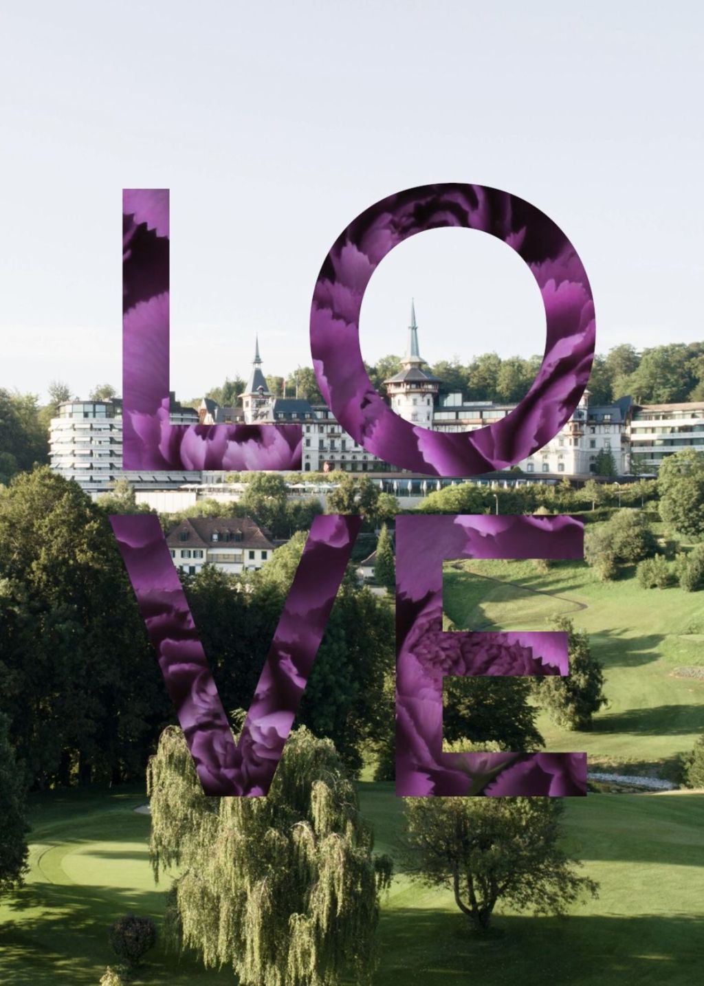 Große lilafarbene Buchstaben mit der Aufschrift "LOVE" sind über eine malerische Landschaft mit grünem Gras, Bäumen und historischen Gebäuden mit Türmen im Hintergrund gelegt. Die Buchstaben haben eine florale Textur.