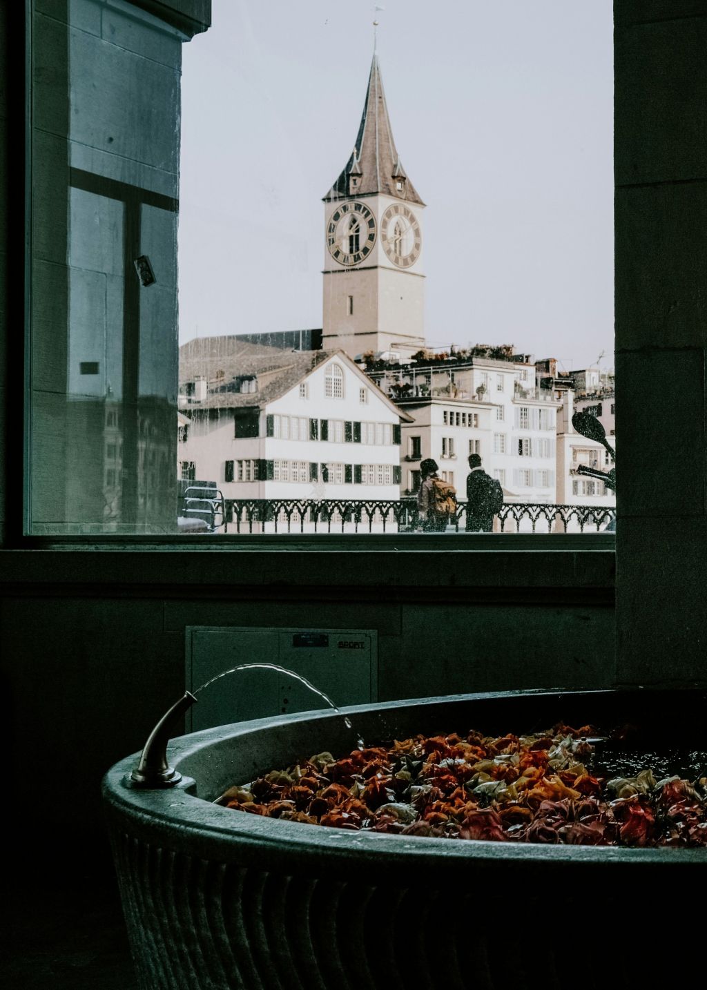 Ein großer steinerner Rosenbrunnen, der mit bunten Blumen gefüllt ist, steht in einem Innenraum in der Nähe eines Fensters, das den Blick auf einen historischen Uhrenturm und alte Gebäude freigibt. Zwei Personen gehen draußen auf der Straße entlang.