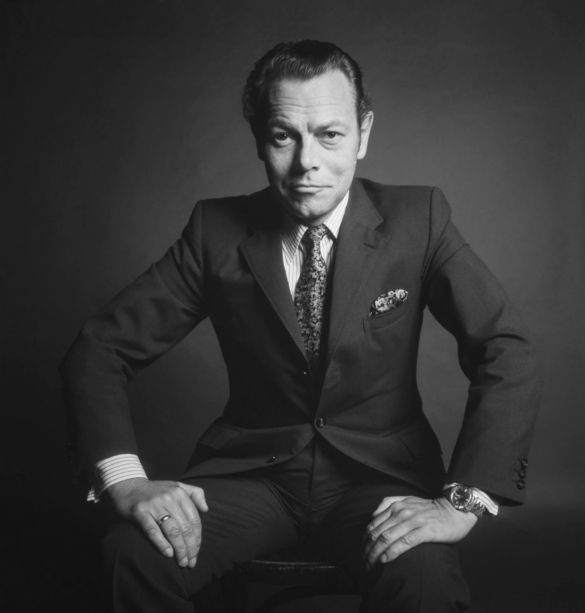 Ein Mann in dunklem Anzug und Krawatte sitzt selbstbewusst auf einem Hocker vor einem schlichten dunklen Hintergrund und blickt mit einem leichten Lächeln und auf die Knie gestützten Händen direkt in die Kamera.
