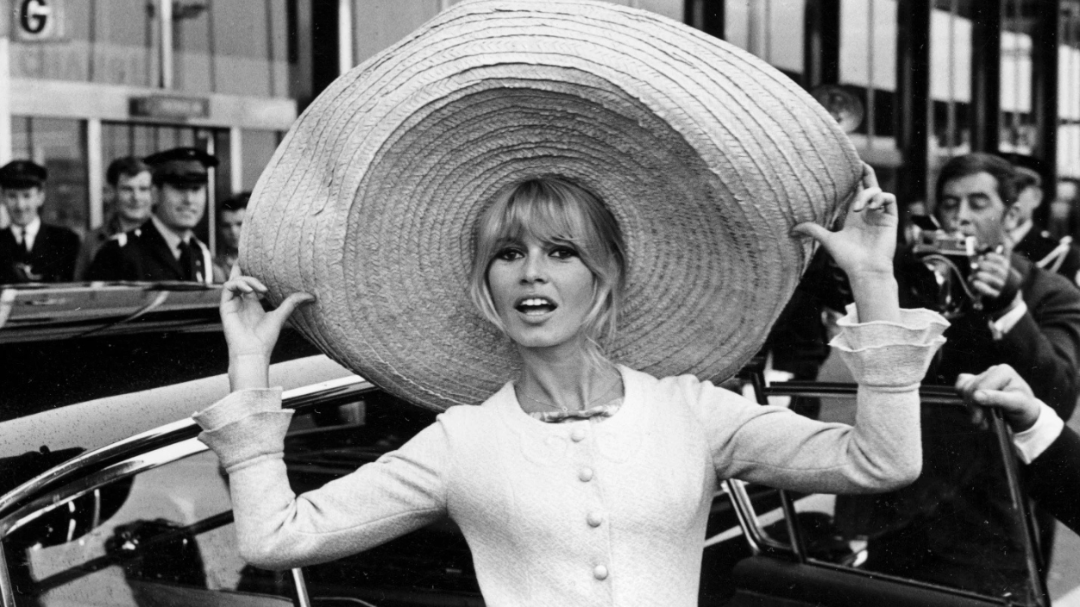 Eine Frau, die einen großen, breitkrempigen Hut und ein geknöpftes Kleid trägt, steigt aus einem Auto und lächelt, während die Leute im Hintergrund zusehen und Fotos machen.