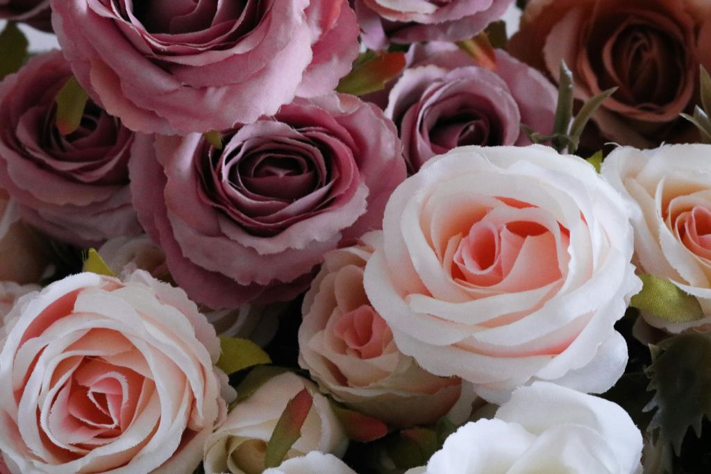 Eine Nahaufnahme mehrerer künstlicher Rosen in zarten Rosa-, Creme- und Mauvetönen, mit Schichten von zarten Blütenblättern und einigen grünen Blättern, die zwischen den Blumen sichtbar sind.