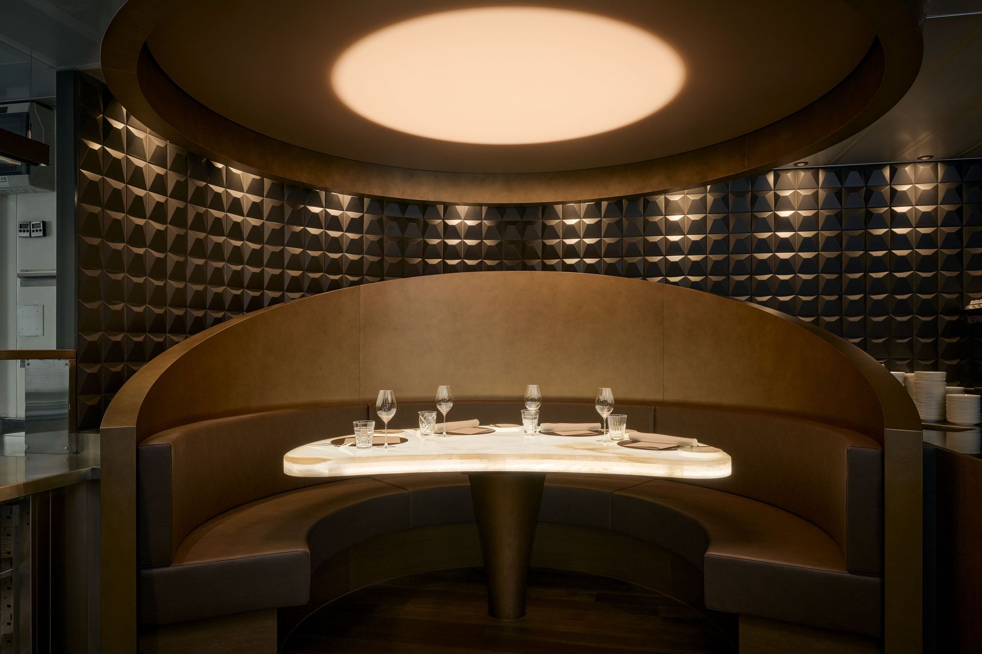 Eine moderne Restaurantkabine im The Restaurant, einem mit 2 Michelin-Sternen ausgezeichneten Restaurant in Zürich, mit einer geschwungenen Lederbank, einem leuchtenden ovalen Tisch für vier Personen und dunklen geometrischen Wandpaneelen unter einer großen runden Deckenleuchte.