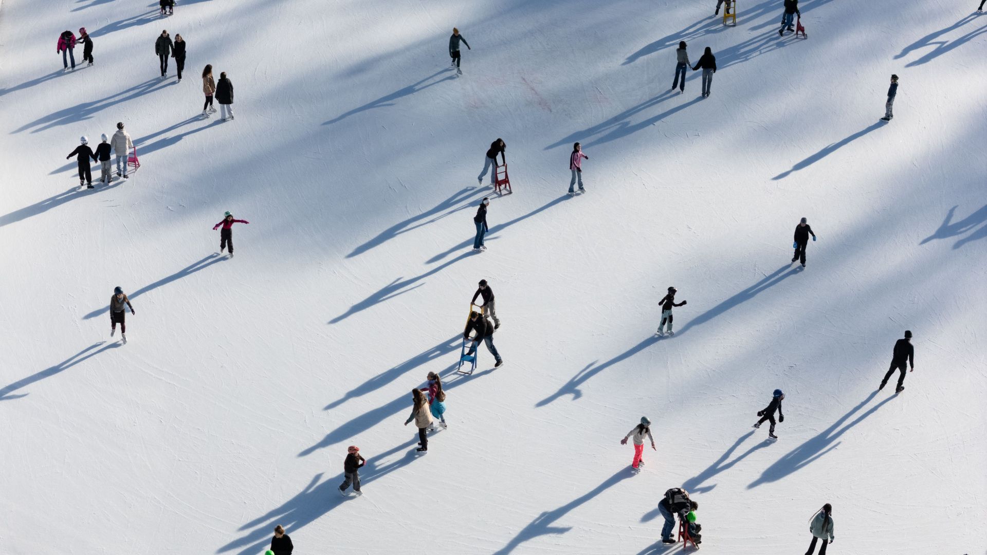 Luftaufnahme von Schlittschuhläufern auf einer Eisbahn während der familien Ferien, die lange Schatten auf die weiße Eisfläche werfen. Die Schlittschuhläufer, einige mit Stützen, verteilen sich - perfekt für Reisen mit Kindern in einer malerischen Winterlandschaft.