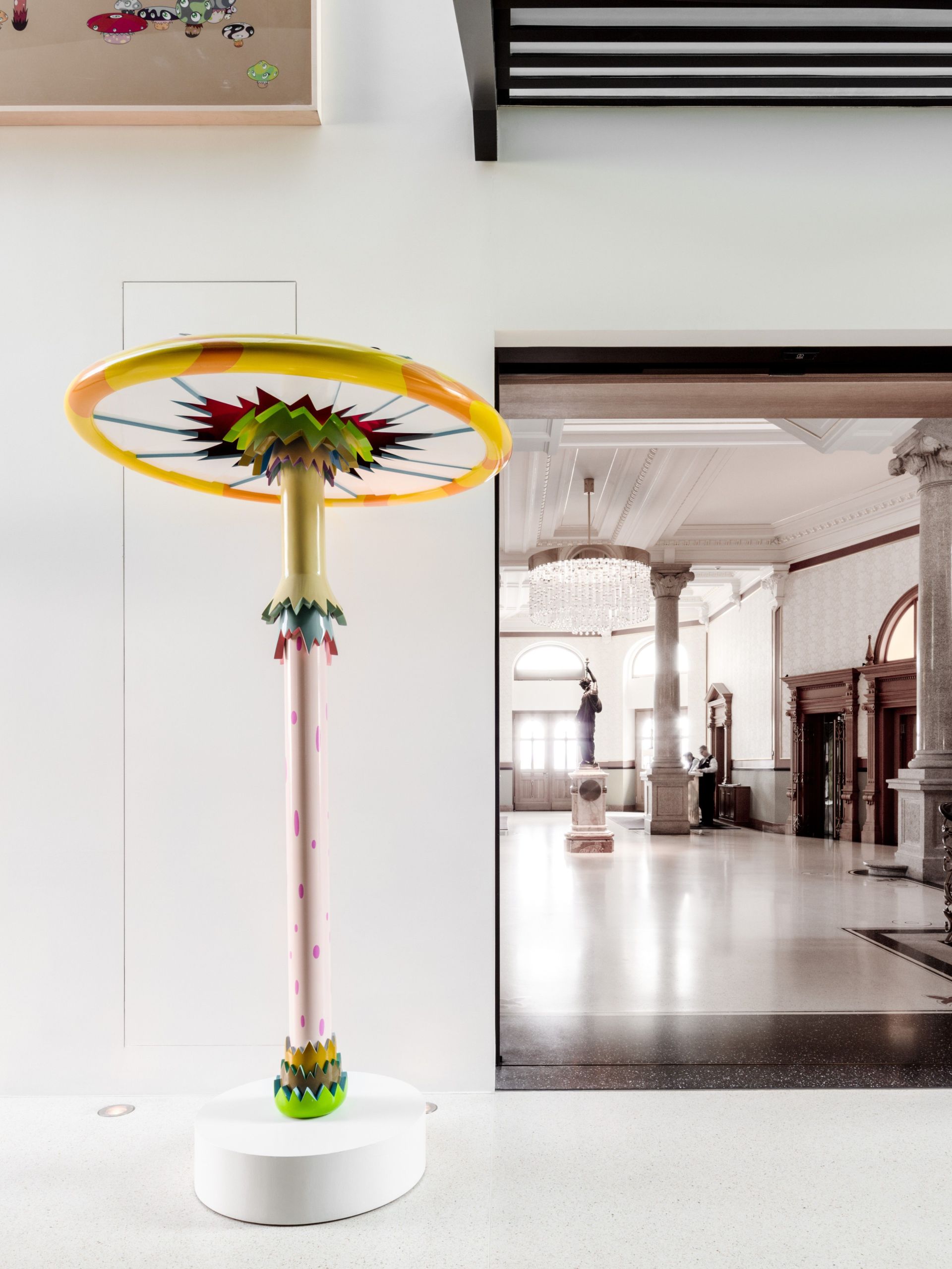 Ein farbenfrohes, verspieltes Skulpturwerk, das einer stilisierten Pflanze oder einem Baum ähnelt, steht auf einem weißen Sockel in einem hellen, modernen Galerieraum und zelebriert die harmonische Verschmelzung von Kunst und Architektur vor dem Hintergrund klassischer Details.