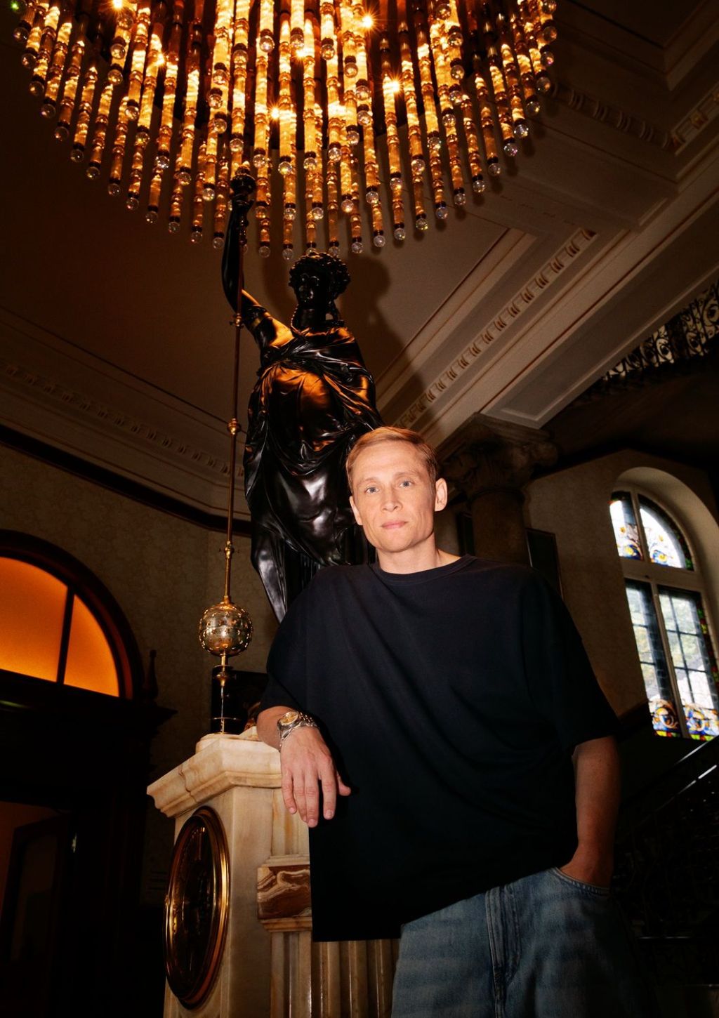 Eine Person in einem dunklen Hemd lehnt an einem Marmengeländer in einem prunkvollen Gebäude mit einem großen Kronleuchter, Buntglasfenstern und einer dunklen Statue im Hintergrund.
