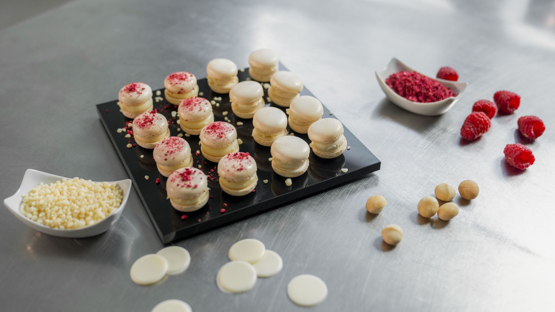 Kleine weiße Macarons, von denen einige mit roten Streuseln verziert sind, liegen auf einem schwarzen Tablett. Um sie herum stehen auf einer Metallfläche Schälchen mit weißen Schokoladenstückchen, zerkleinerten roten Streuseln, frischen Himbeeren und Macadamia-Nüssen.