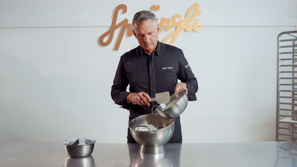 Ein Koch in einer dunklen Uniform gießt Teig aus einer Metallschüssel in eine andere in einer hellen Küche, hinter ihm steht an der Wand der Schriftzug „Springli“ und rechts davon steht ein Gestell.