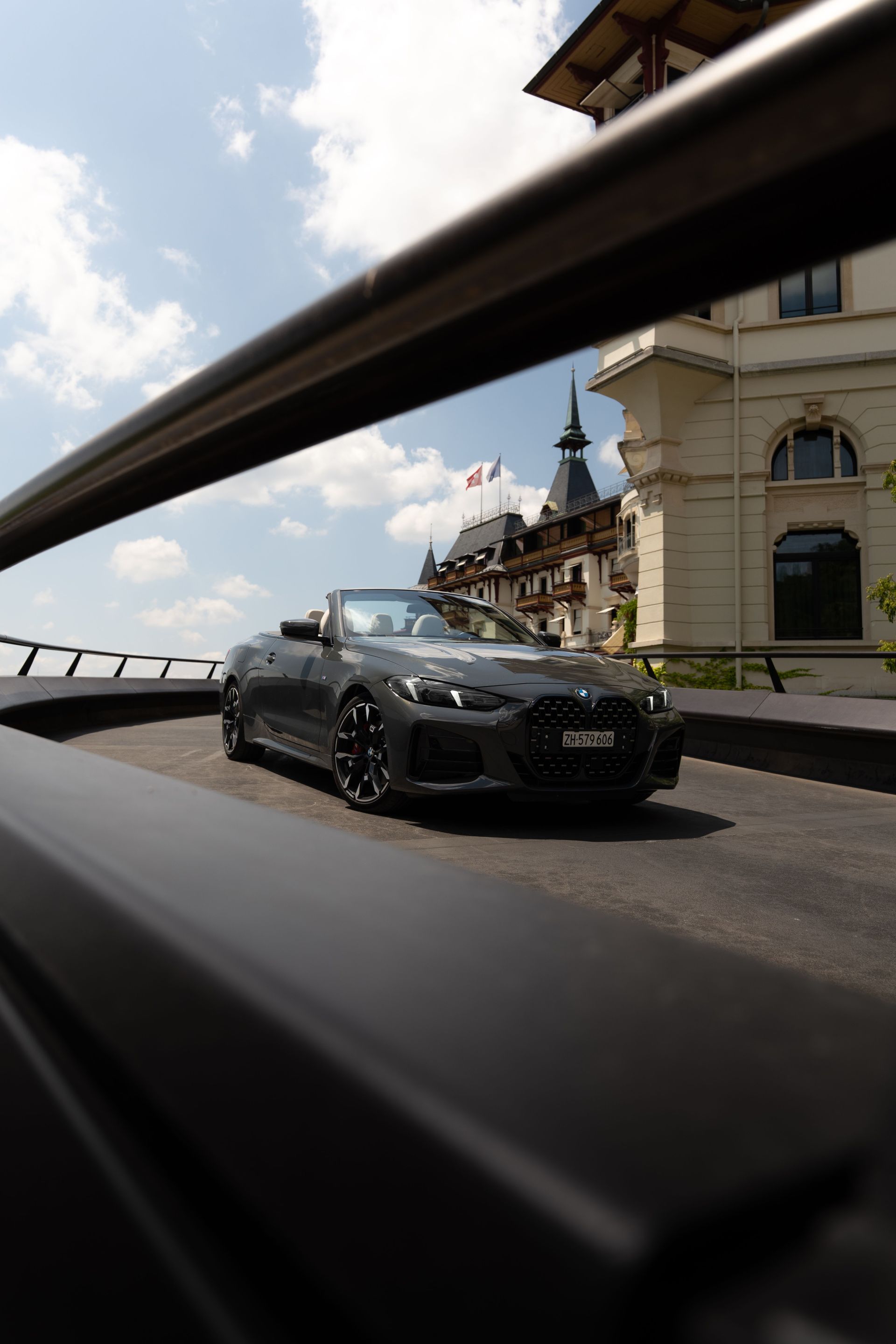 A sleek black convertible car drives on a curved road, framed by metal railings, with a historic building—reminiscent of the renowned Concierge Dolder Grand—and blue sky with clouds in the background.