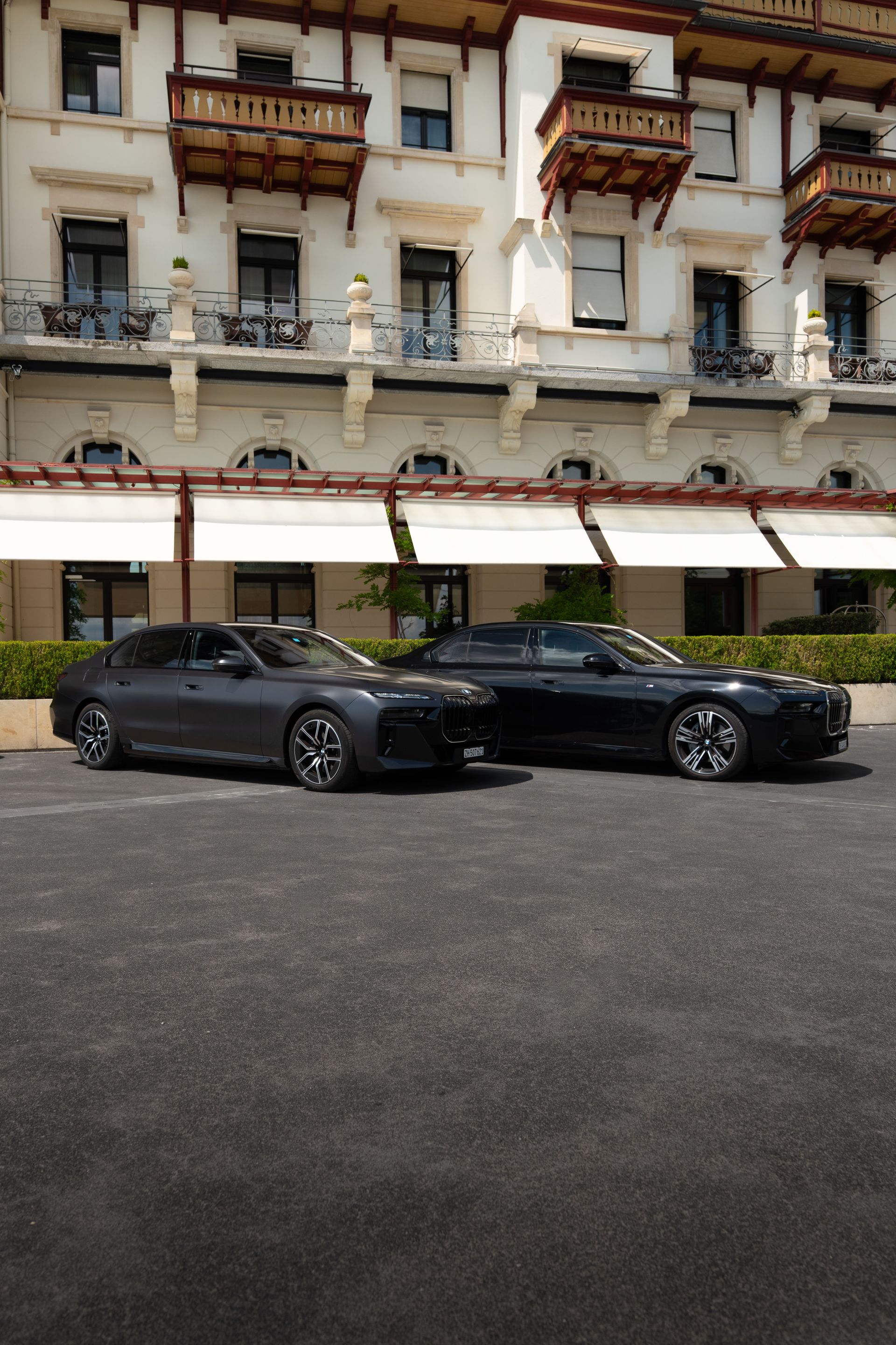 Two sleek, dark-colored cars are parked side by side in front of a grand building with ornate balconies, large windows, and white walls. The elegant scene evokes the refined ambiance of a Concierge Service Zurich, where every detail reflects luxurious comfort and modern sophistication.