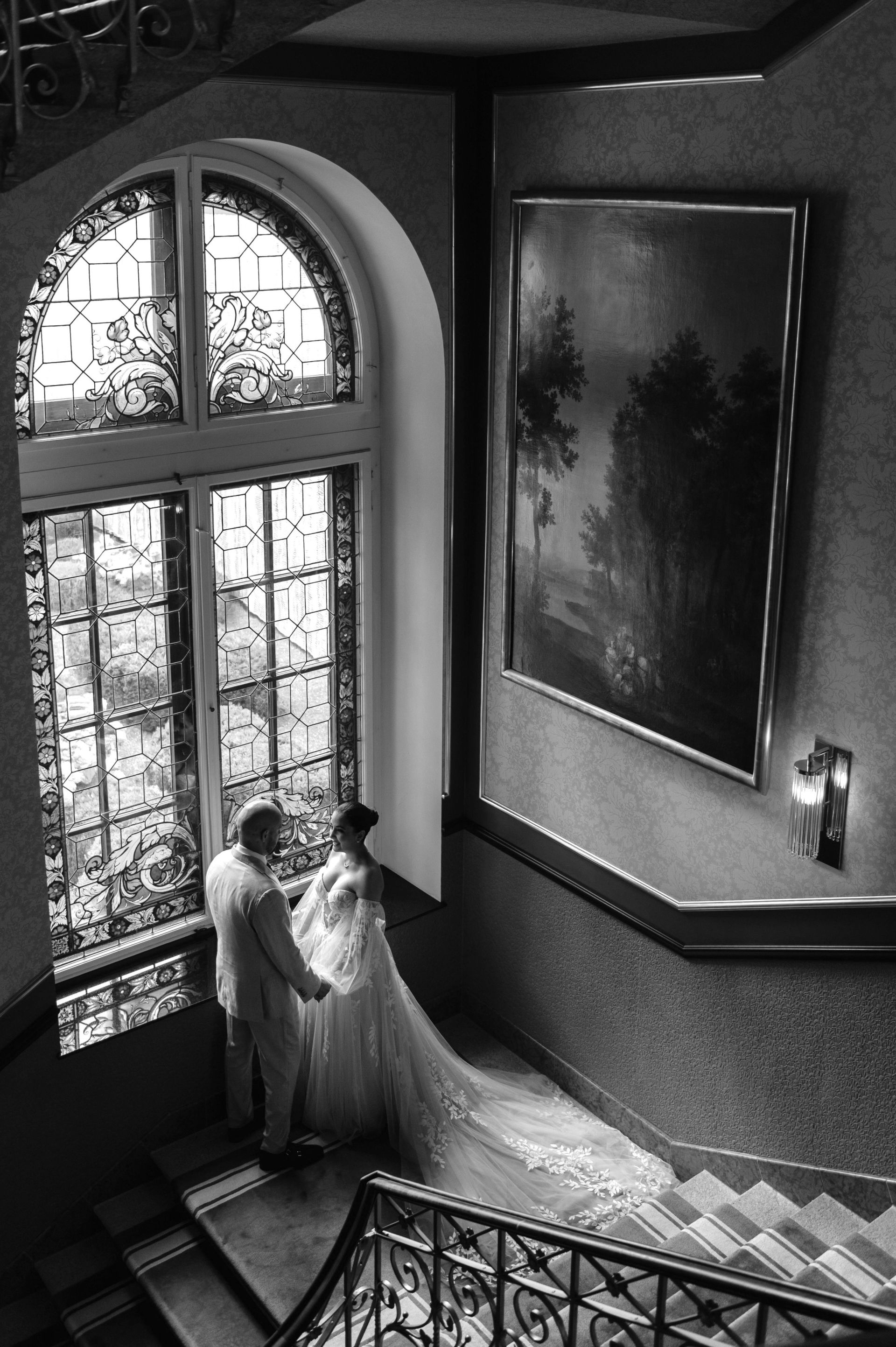 A bride and groom stand holding hands on a staircase in front of a large stained glass window, with a framed painting and elegant railings visible—a timeless moment from a wedding at the Dolder Grand. The scene is captured in black and white.