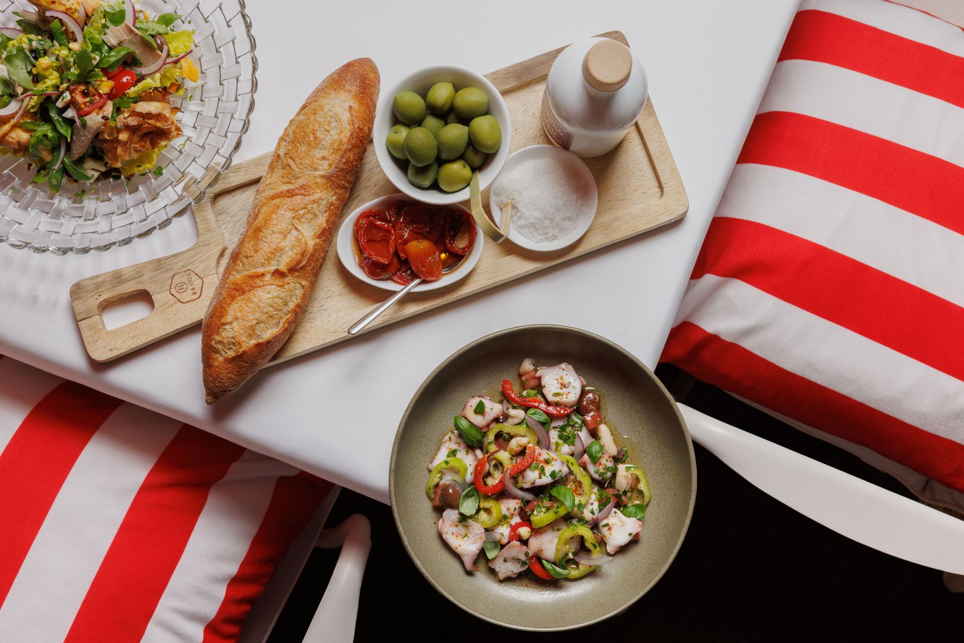 Ein Tisch mit einem Salat, einer Schüssel Ceviche, einem Baguette, grünen Oliven, sonnengetrockneten Tomaten, Salz und Öl auf einem Holztablett. Auf den Stühlen liegen rot-weiß gestreifte Kissen.