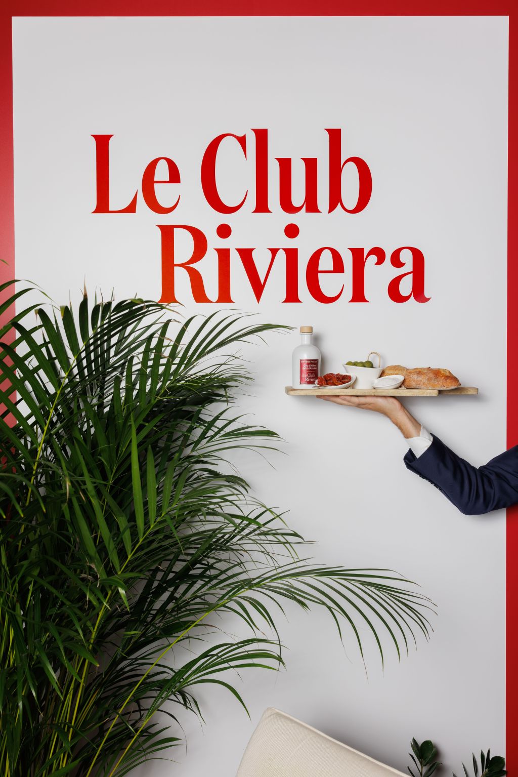 Eine Hand hält ein Tablett mit einem Getränk, Brot und Snacks vor einer weißen Wand, auf der in roter Schrift „Le Club Riviera“ steht. Außerdem sind grüne Blätter und ein Teil eines Sofas zu sehen.