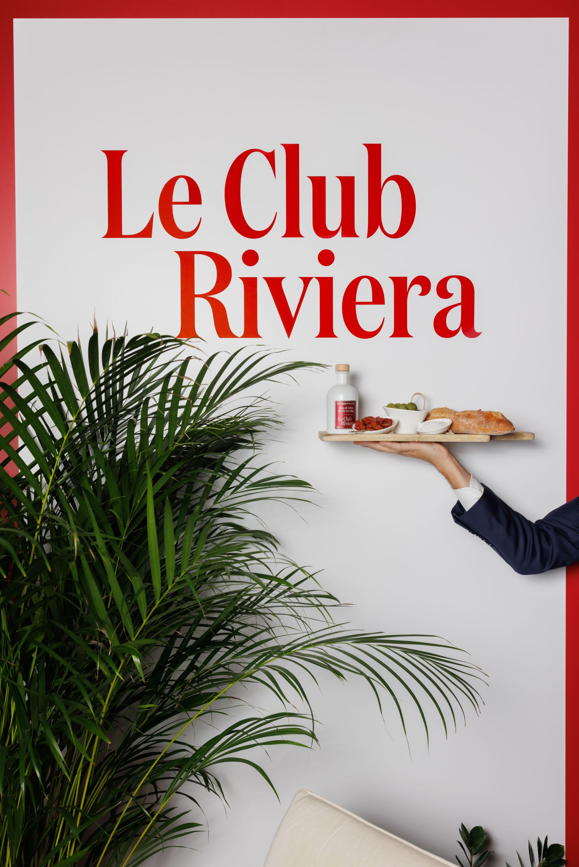 Eine Hand hält ein Tablett mit einem Getränk, Brot und Snacks vor einer weißen Wand, auf der in roter Schrift „Le Club Riviera“ steht. Außerdem sind grüne Blätter und ein Teil eines Sofas zu sehen.
