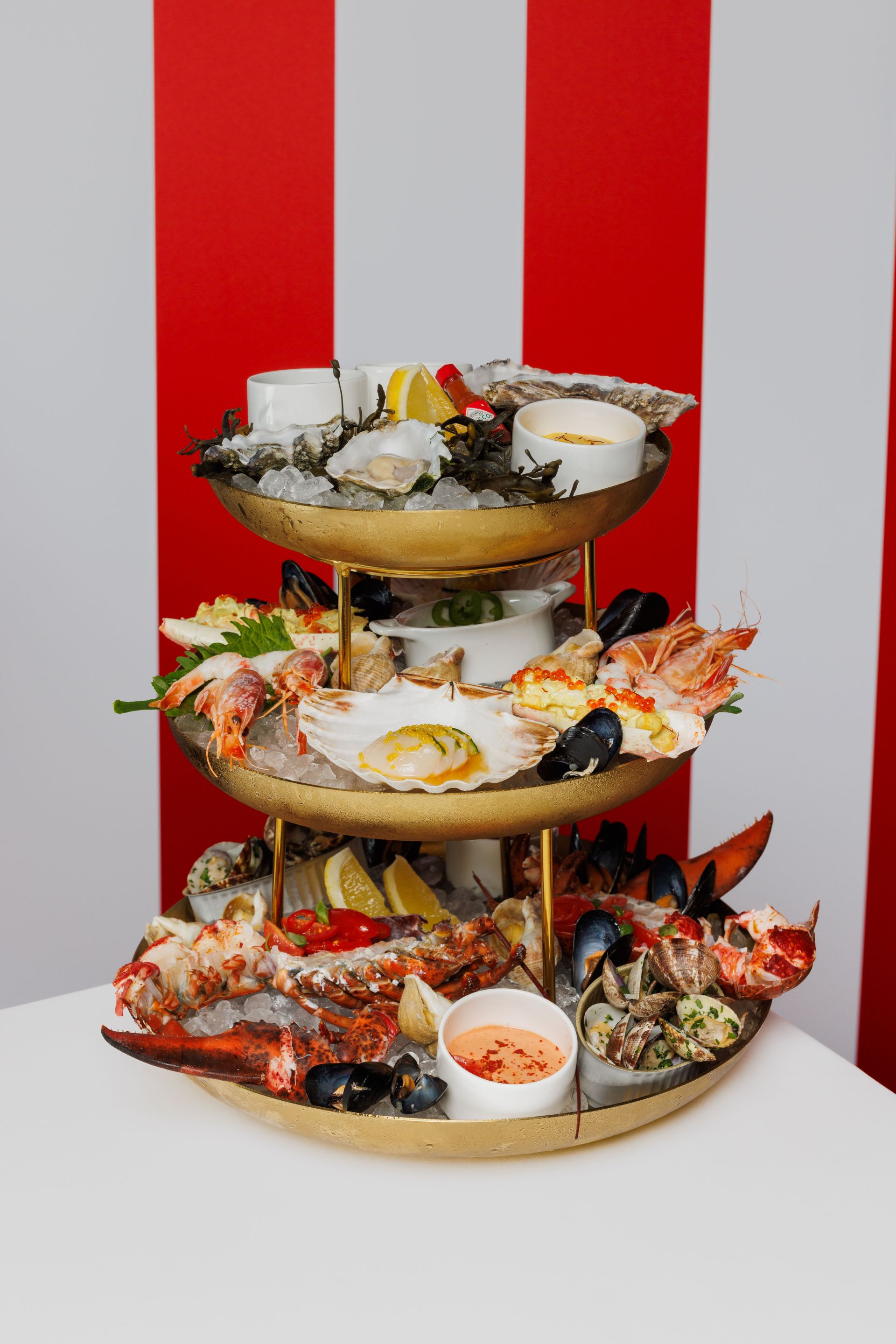 Eine dreistöckige Meeresfrüchteplatte mit Garnelen, Austern, Miesmuscheln, Hummer, Zitronen und Dip-Saucen vor einem rot-weiß gestreiften Hintergrund.