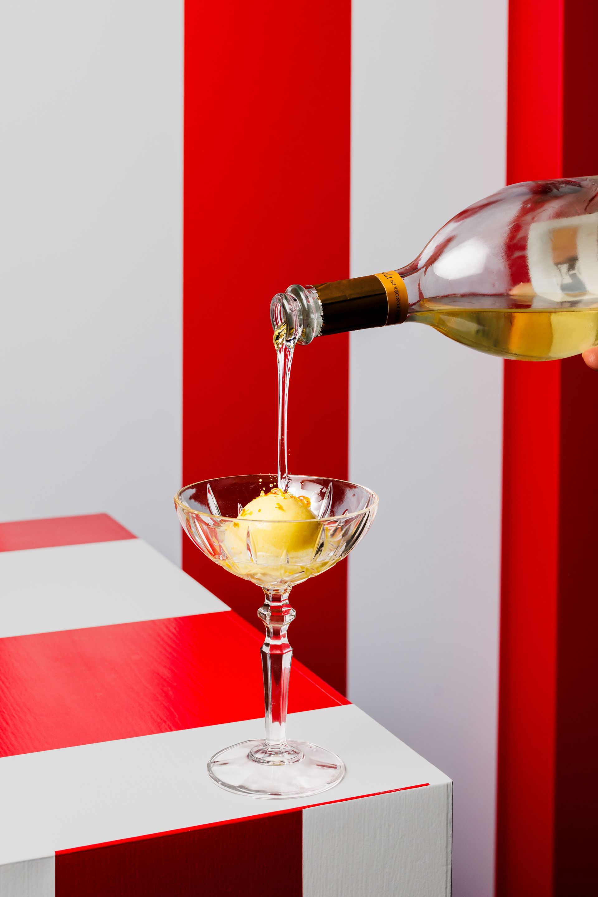 Eine Hand gießt Weißwein über eine Kugel Sorbet in einem Glas, vor einem rot-weiß gestreiften Hintergrund und einem rot-weiß gestreiften Tisch.