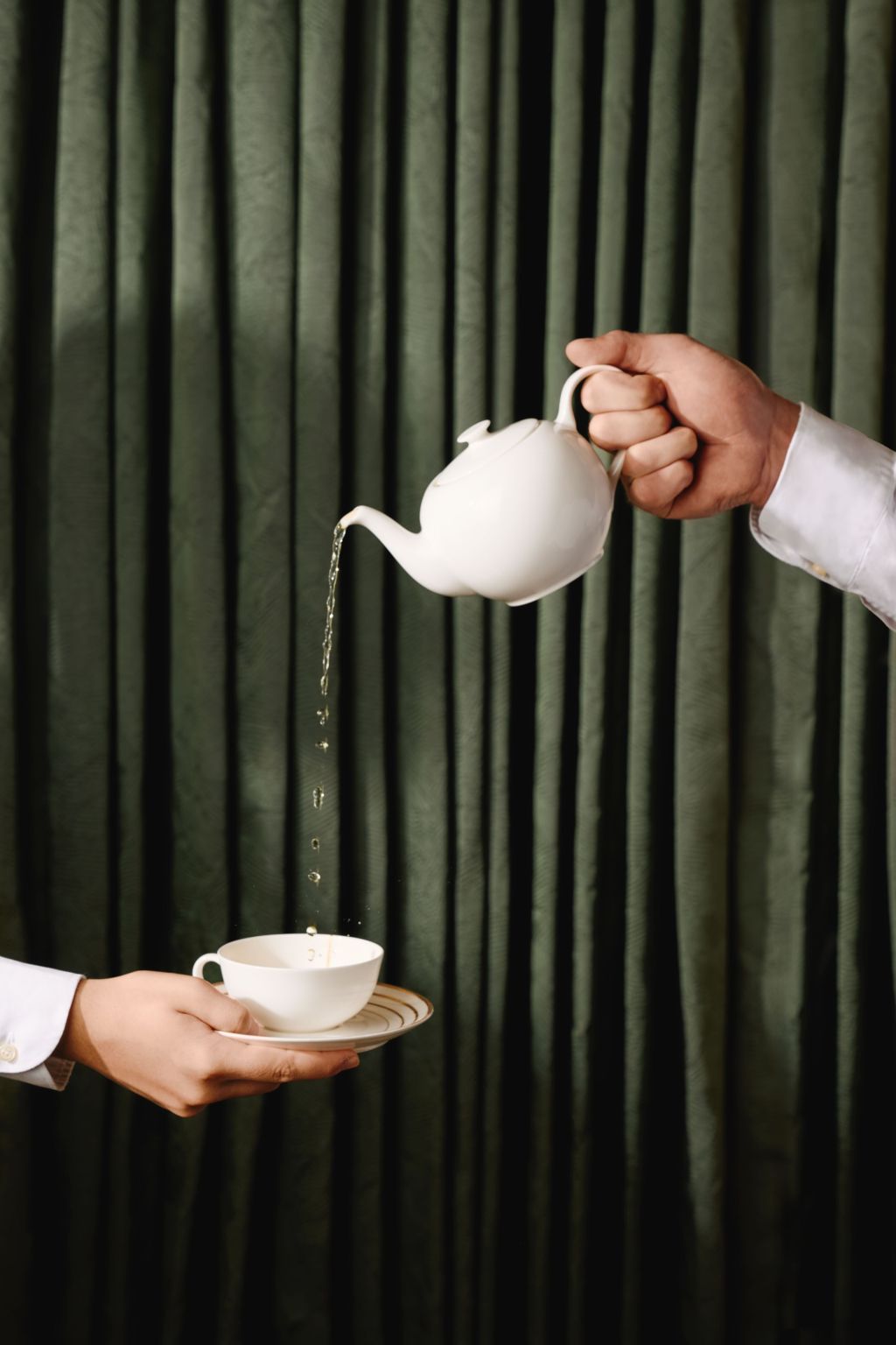Vor einem grünen Vorhang gießt eine Person Tee aus einer weißen Teekanne in eine weiße Teetasse, die von einer anderen Person gehalten wird.