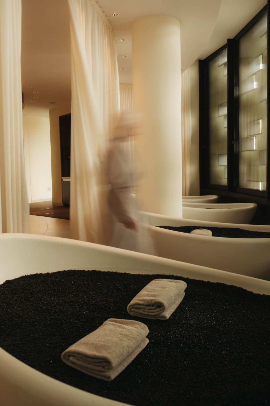Weiße Badewannen, gefüllt mit schwarzen Kieselsteinen und gefalteten Handtüchern, in einem Dolder Grand Spa-Raum, durch den eine verschwommene Gestalt vorbeigeht.